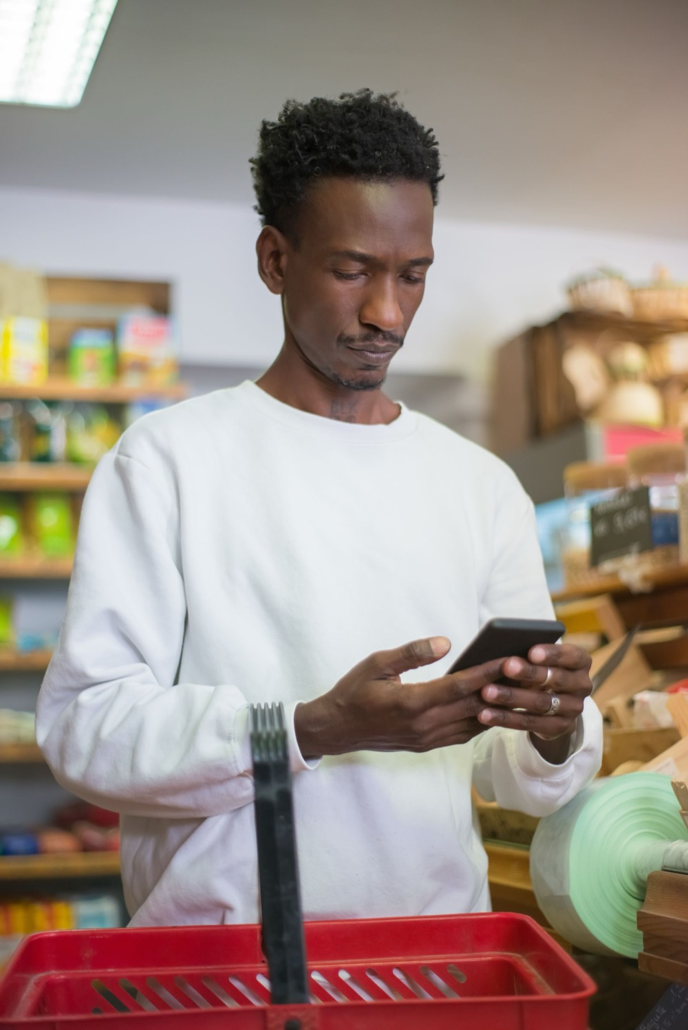 Homem olhando o celular com uma cesta de compras numa loja